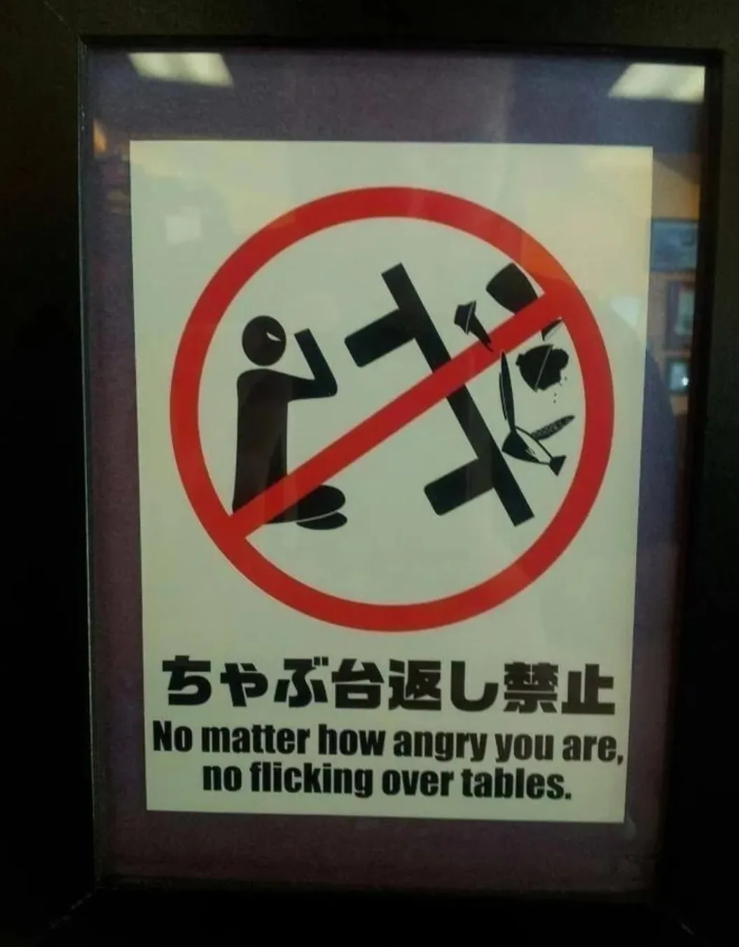 A framed Japanese sign with a red prohibition circle over a black pictogram of a person flipping a table. Below, Japanese…