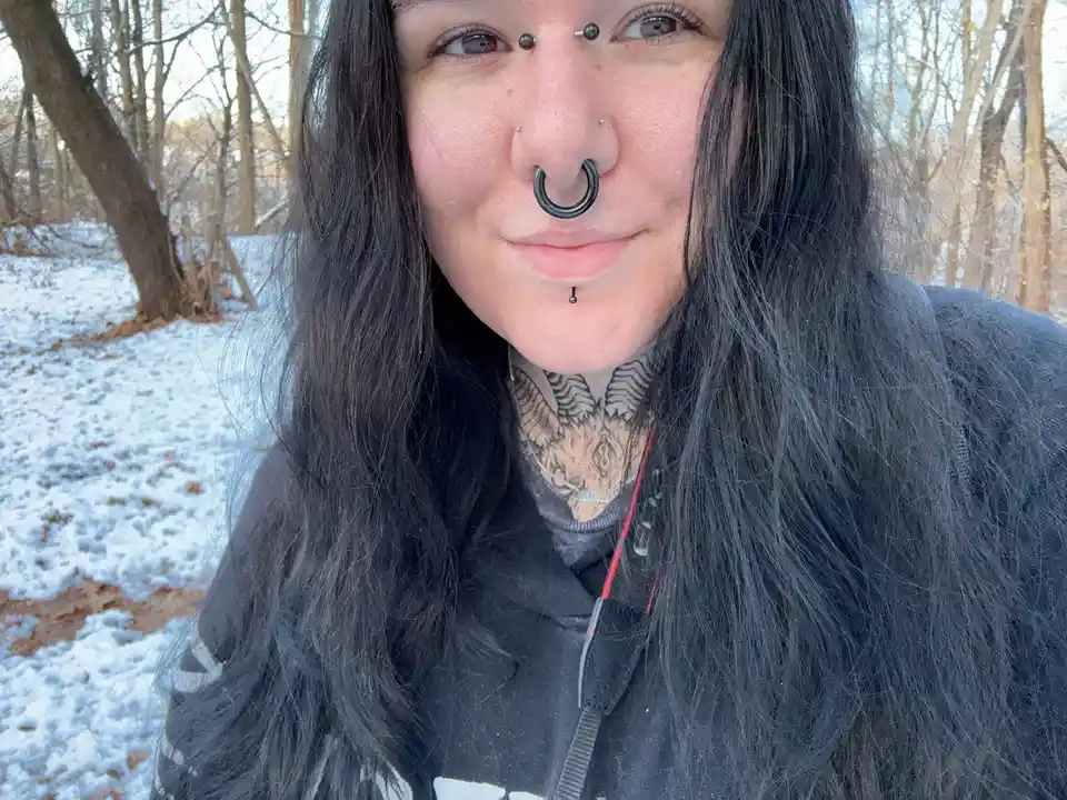 A person with long dark hair and multiple facial piercings, including a large black nose ring, stands outdoors in a snowy…