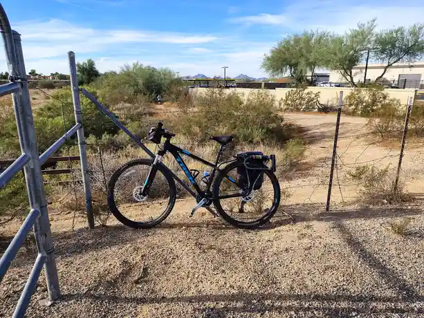 A black Trek mountain bike with blue accents and panniers is leaning against a metal fence gate in a dry, gravelly desert…