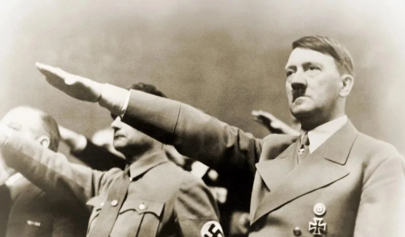 A black-and-white historical photograph shows Adolf Hitler, wearing a Nazi uniform with a swastika armband and Iron Cross…