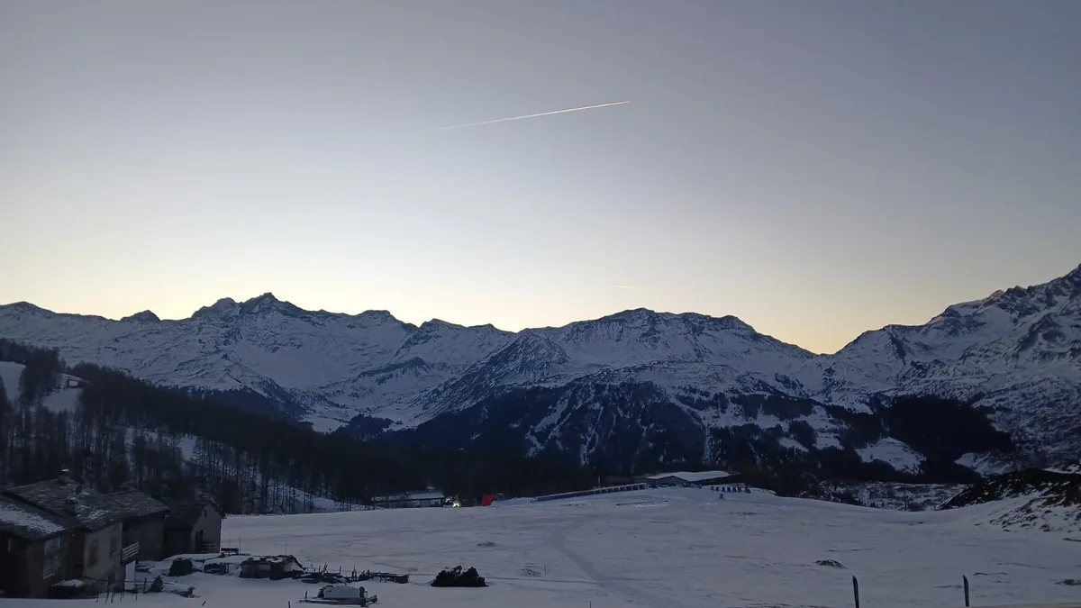 A serene snowy mountain landscape at dusk with a long airplane contrail visible in the pale sky. The foreground shows…