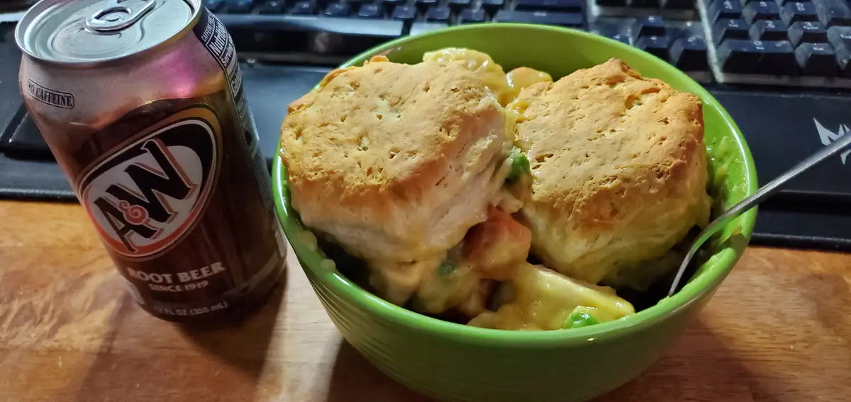 A green bowl filled with a creamy casserole topped with two golden-brown biscuits sits on a wooden desk next to a can of A&W…