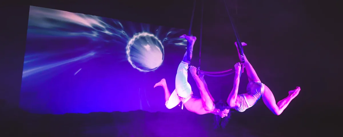 Two acrobats hang upside down from ropes in a dark, purple-lit stage, performing a synchronized aerial act. Behind them, a…