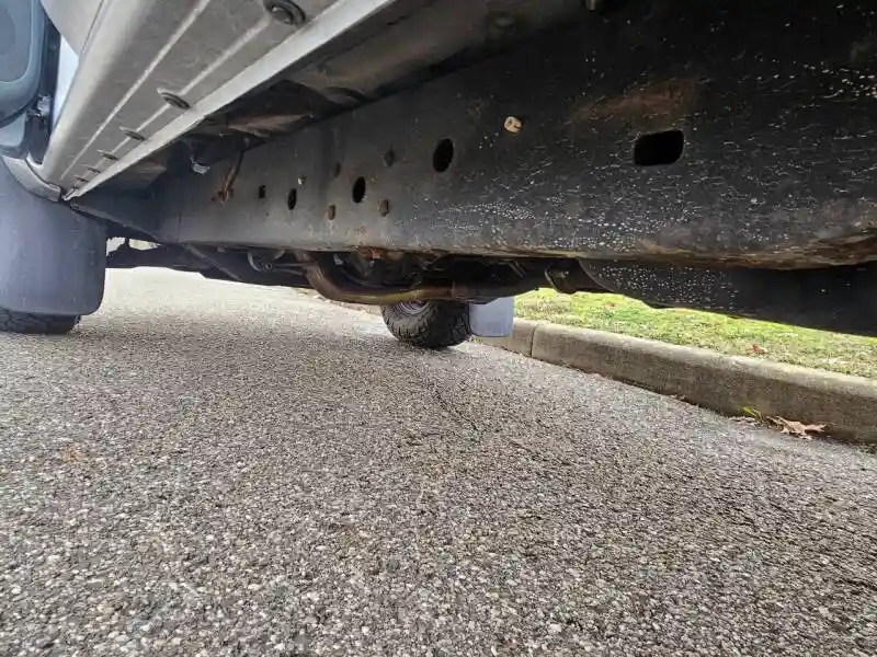 A low-angle photo shows the underside of a vehicle, revealing a rusty, bolted metal frame, exhaust pipe, and a tire…