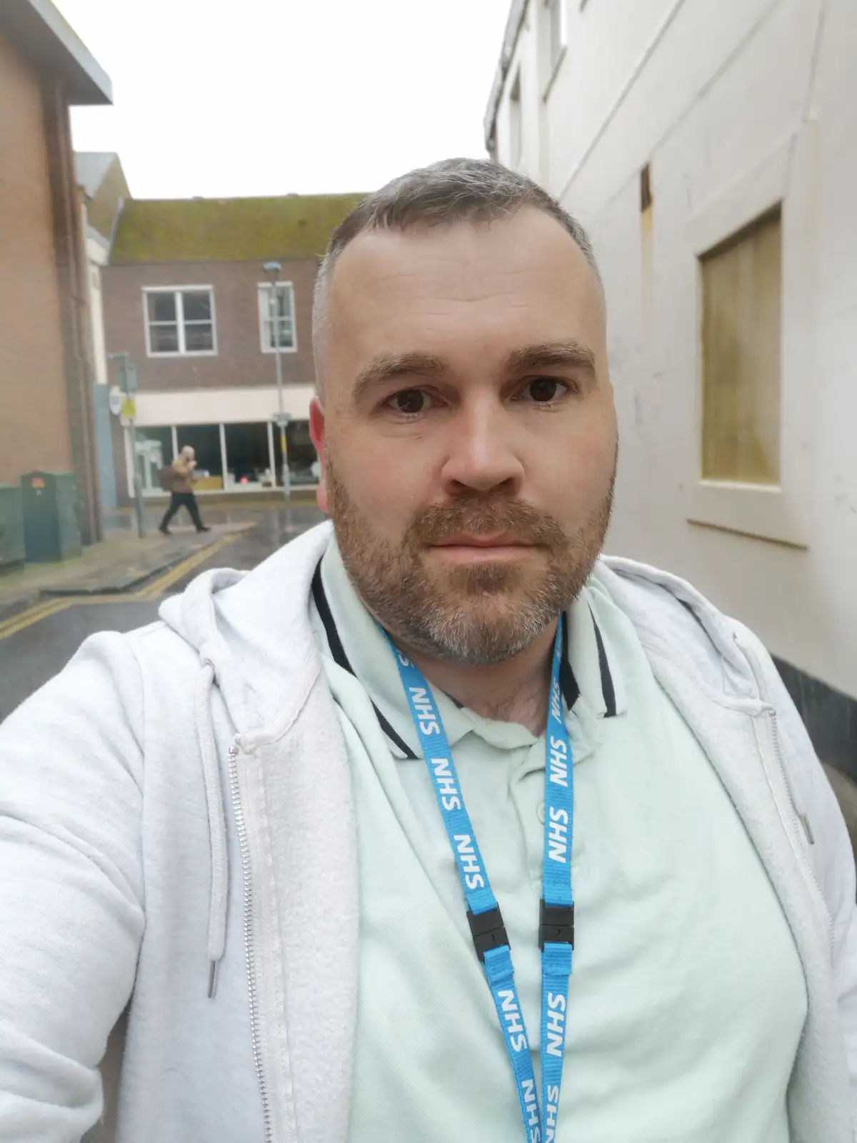 A man with a short beard and gray hair takes a selfie outdoors, wearing a light green polo shirt under a white zip-up…