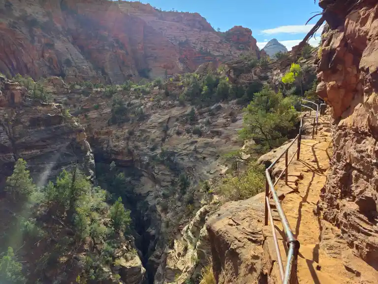 A scenic photo of a narrow, elevated hiking trail with a metal railing, clinging to the side of a steep, red rock canyon…
