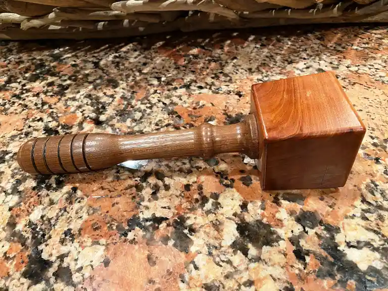 A polished wooden mallet with a square head and ribbed handle rests on a speckled granite countertop, with a stack of…