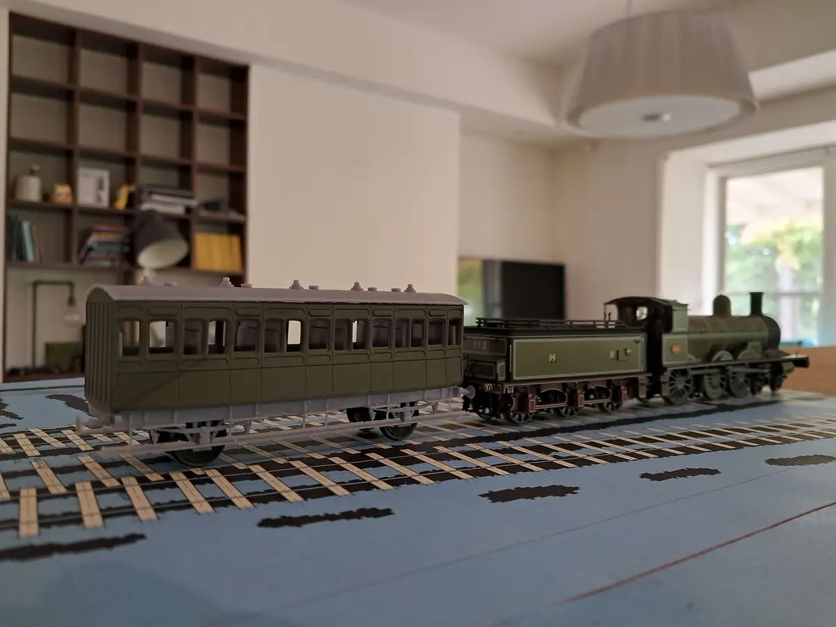 A green model train with a locomotive and passenger car sits on a printed track layout on a blue table, with a blurred…
