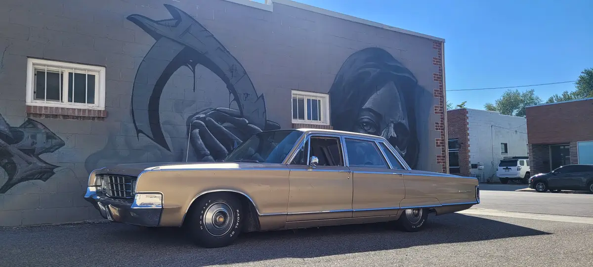 A tan vintage Lincoln Continental sedan is parked in front of a large mural depicting the grim reaper holding a scythe. The…