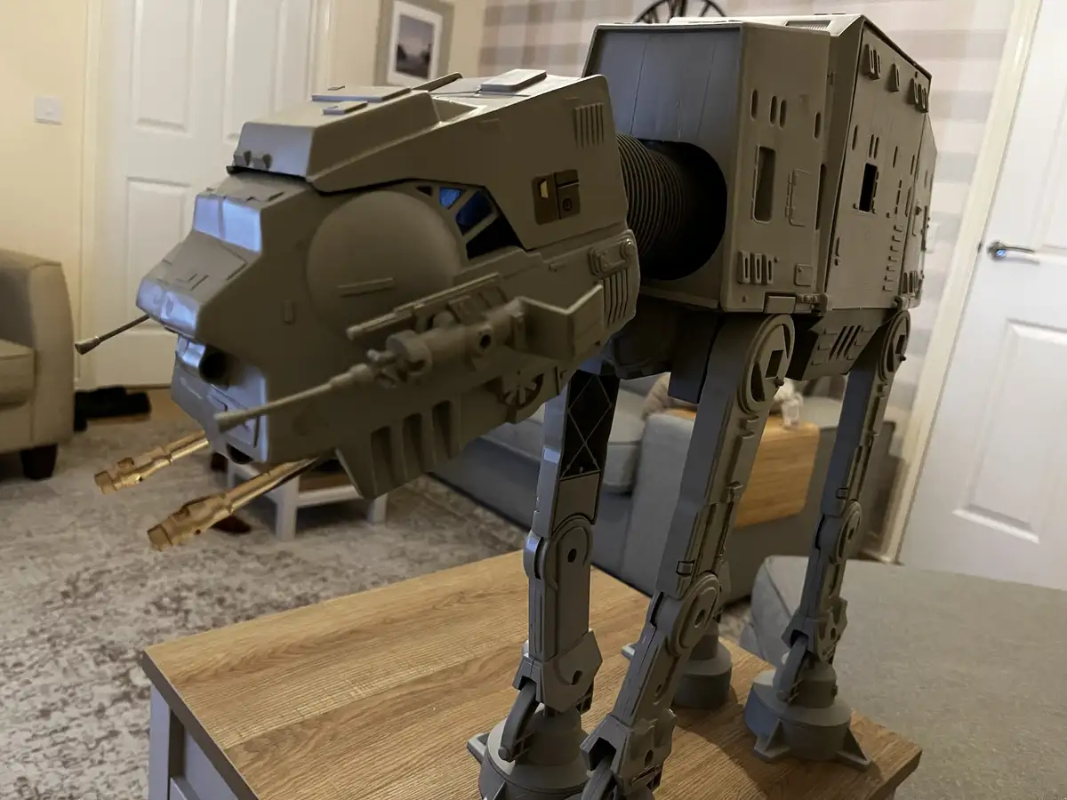 A large, grey plastic AT-AT walker toy from Star Wars is standing on a wooden coffee table in a living room. The toy has its…