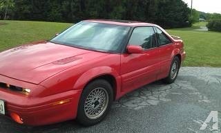 a red 1980s-style sports car, likely a Mazda RX-7, is parked on a cracked asphalt driveway with grass and trees in the…