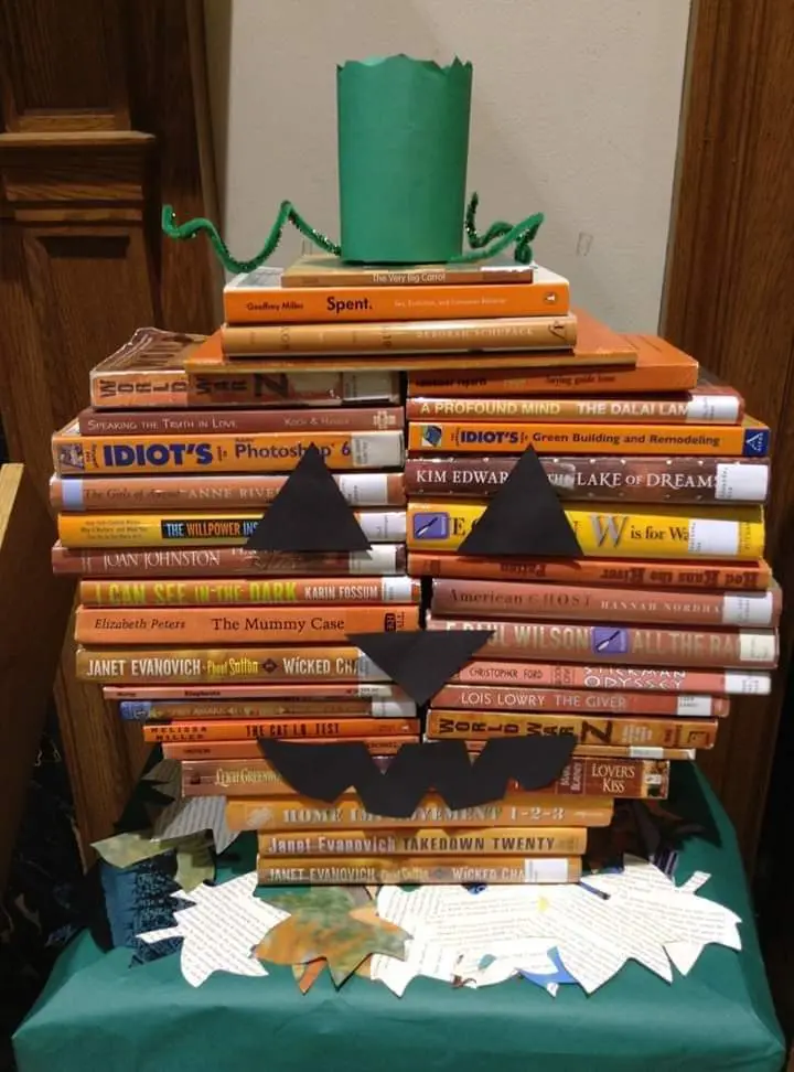 A stack of books is arranged to resemble a jack-o'-lantern, with black paper cutouts forming eyes, a nose, and a mouth, and…