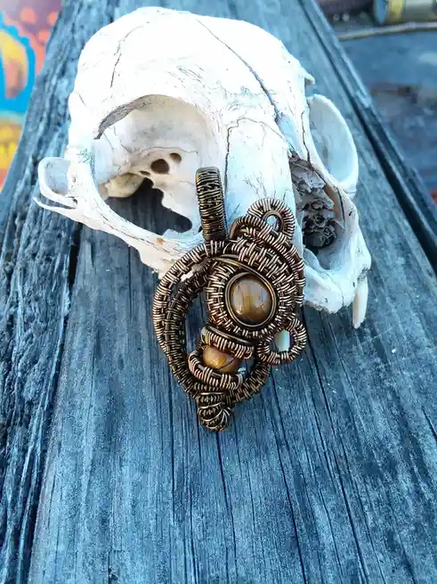 A close-up photo shows a copper wire-wrapped pendant with a tiger's eye stone resting on a white animal skull, which is…