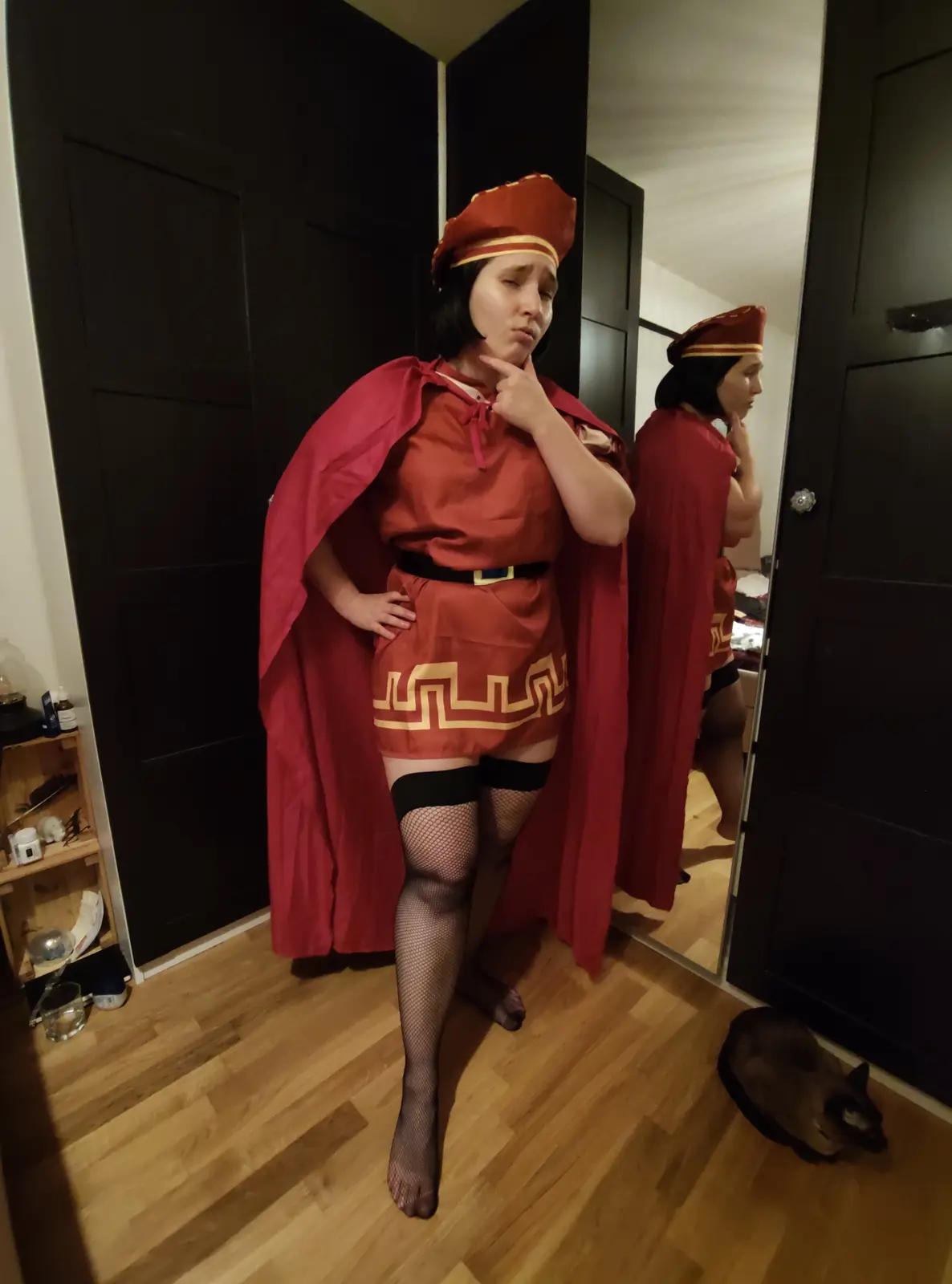 A person in a red and gold Roman-style costume with a cape, fishnet stockings, and thigh-highs stands in front of a mirror…