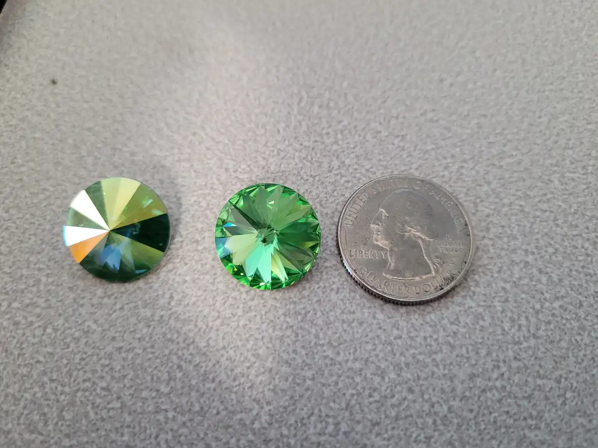 Three round, faceted green crystals are laid side-by-side on a speckled gray surface, with a U.S. quarter dollar coin to the…