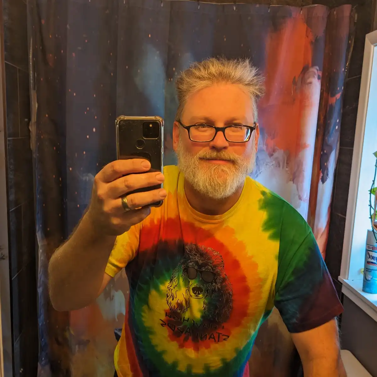 A man with a gray beard and glasses takes a mirror selfie in a bathroom, wearing a colorful tie-dye t-shirt featuring a dog…