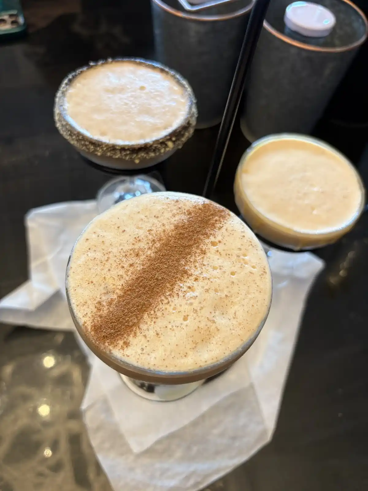 Three creamy, frothy drinks in coupe glasses sit on a dark countertop, one in the foreground topped with a dusting of…