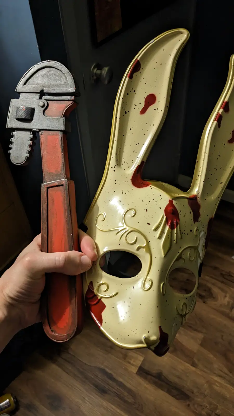 A hand holds a red and silver adjustable wrench next to a cream-colored bunny mask with gold trim and red splatter paint…