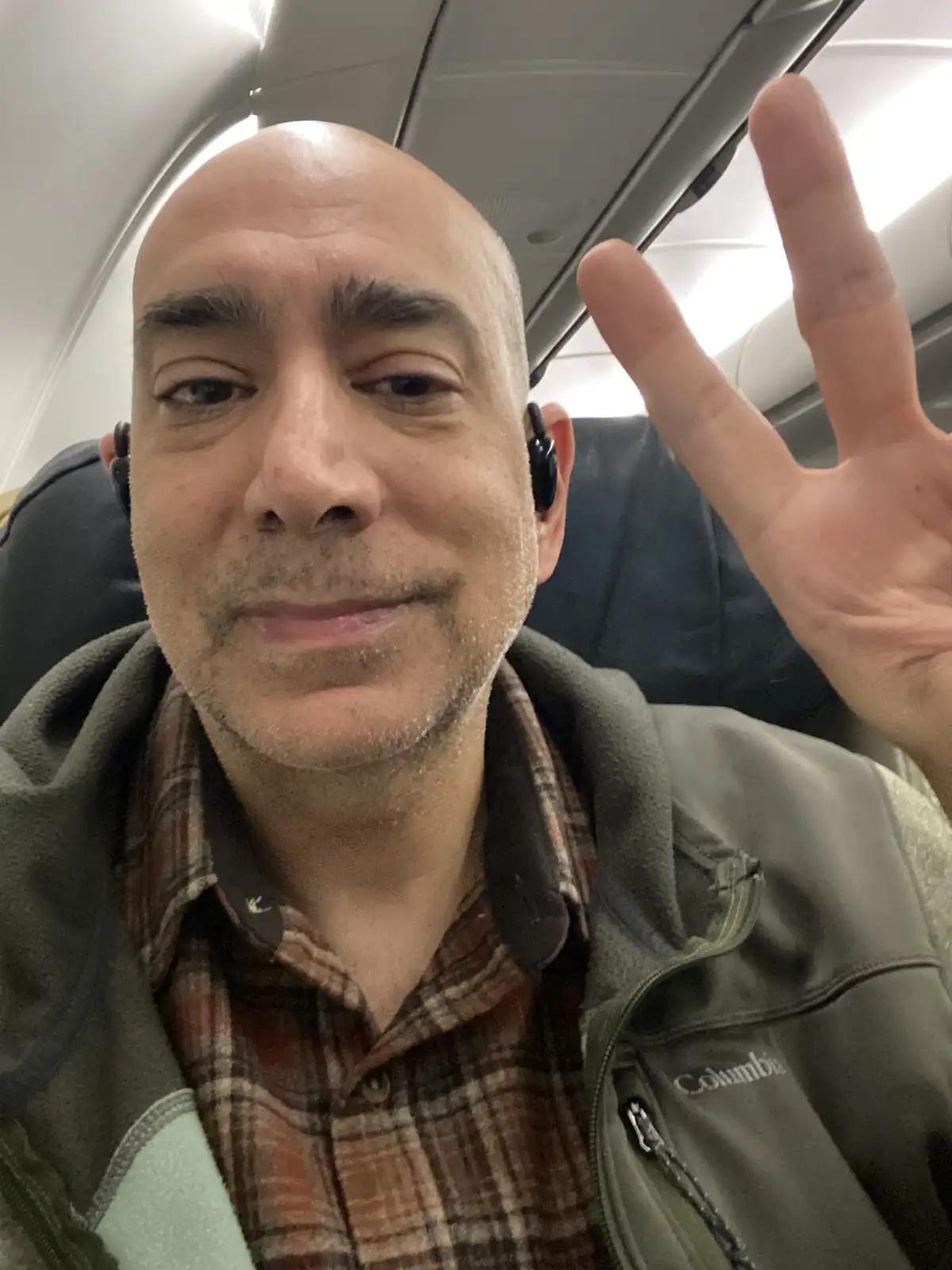 A bald man with a light beard and mustache, wearing a plaid shirt under a green Columbia fleece jacket and black earbuds…