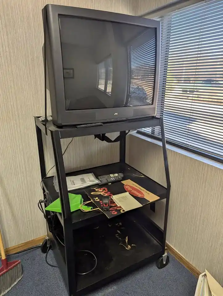 A black JVC CRT television sits on a black rolling cart with two shelves. The top shelf holds only the TV, while the bottom…