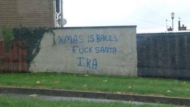 A concrete wall with blue spray-painted graffiti that reads 'XMAS IS BALLS', 'FUCK SANTA', and 'IRA'. The wall is situated…