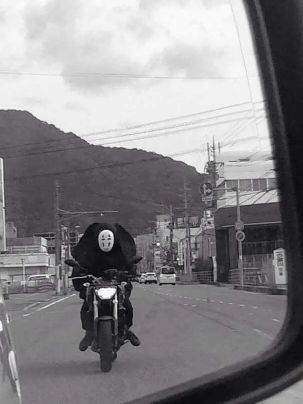 A black-and-white photo taken from inside a car shows a person riding a motorcycle down a street. The rider is wearing a…