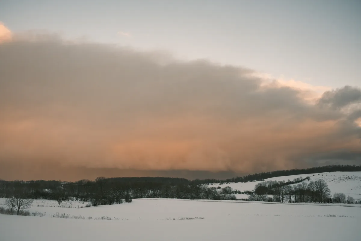 A serene winter landscape with snow-covered fields and bare trees under a dramatic sky with orange and gray clouds. The…