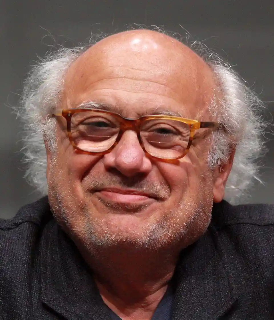 A close-up photo of comedian and actor Danny DeVito, who has a balding head with white hair on the sides, wears…