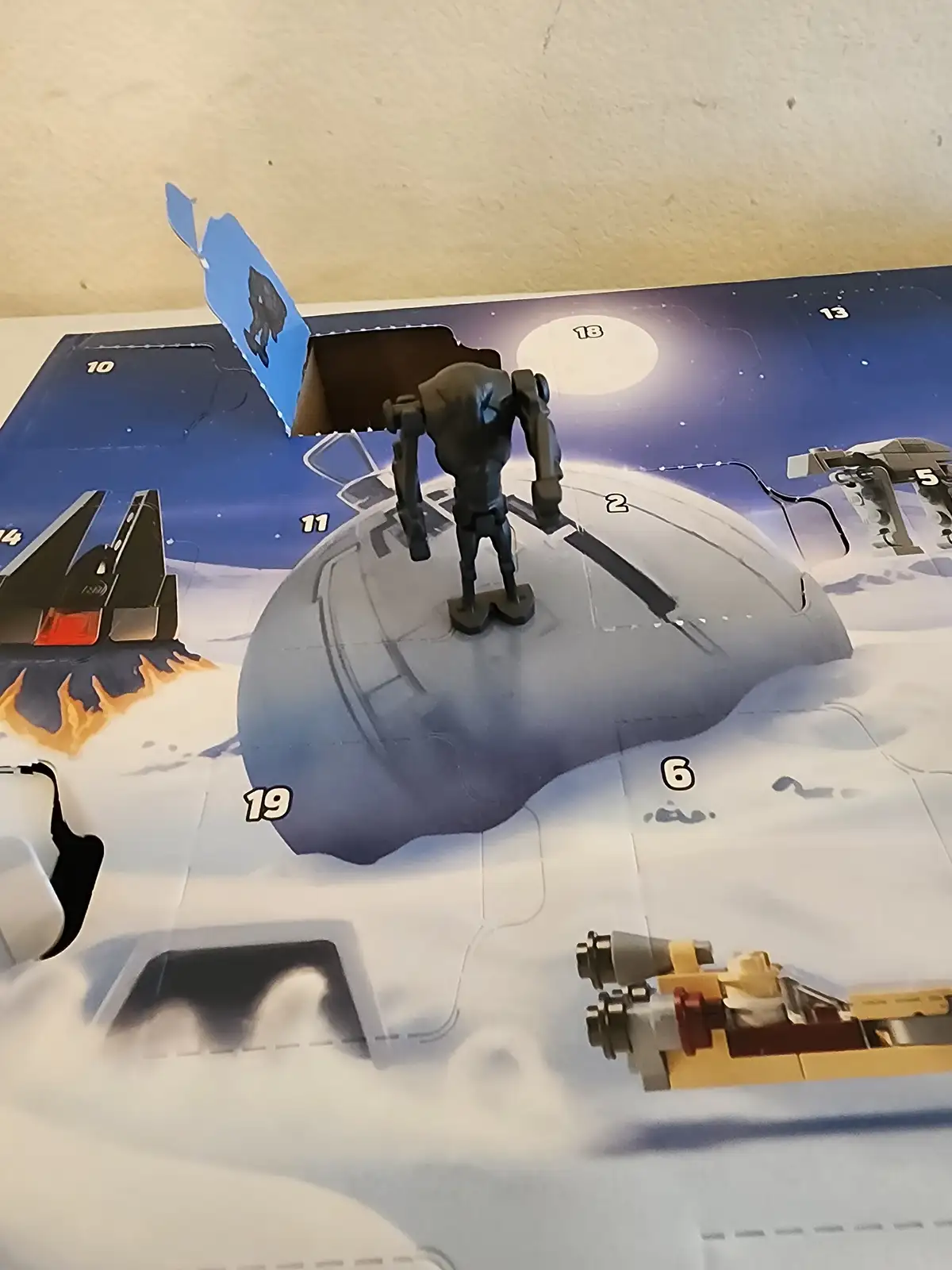 A small black robot toy is placed on a printed LEGO Star Wars advent calendar, standing on a numbered panel. The calendar…