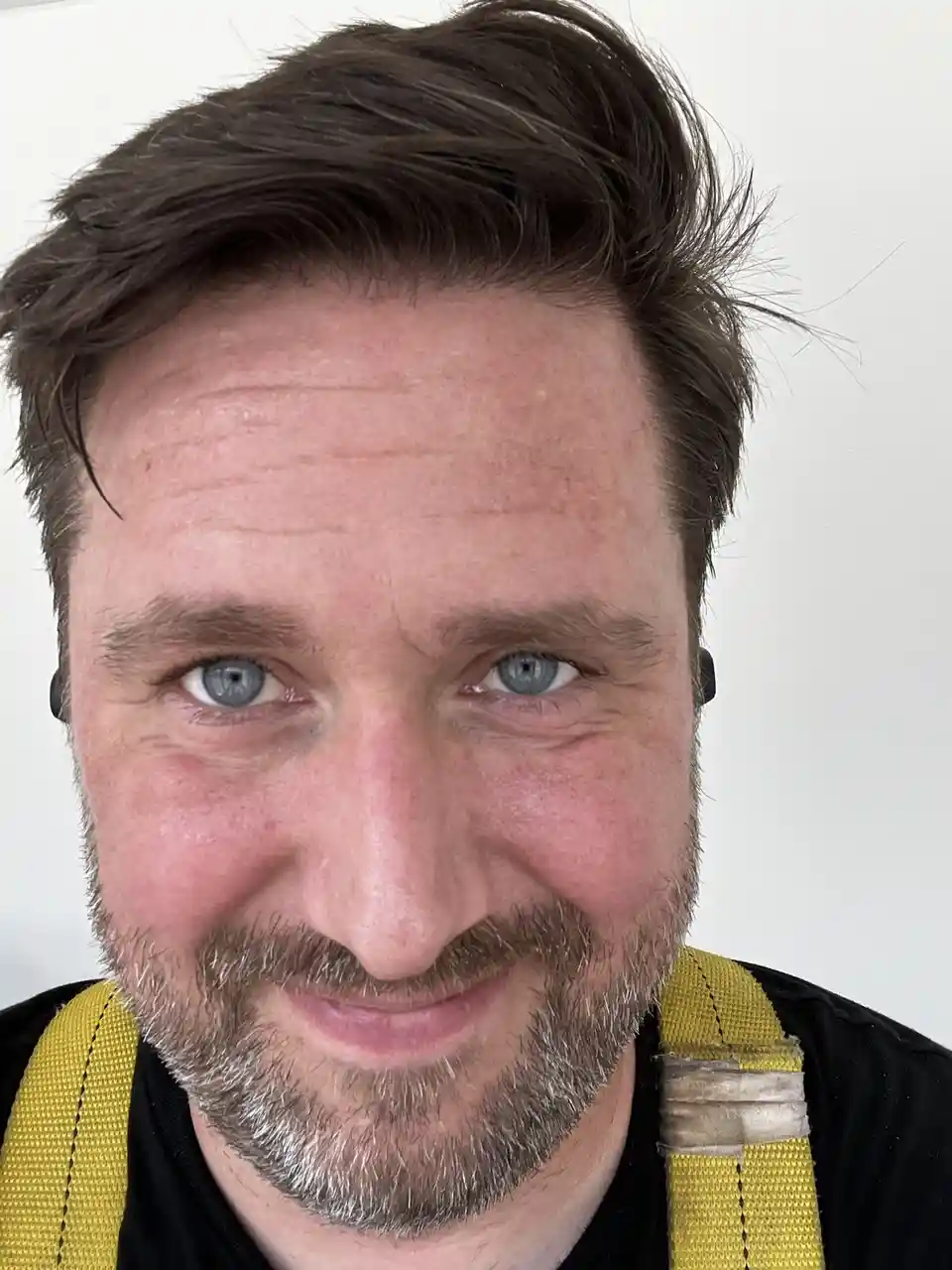 A close-up photo of a man with blue eyes, brown hair, and a graying beard, wearing yellow safety harness straps over a black…