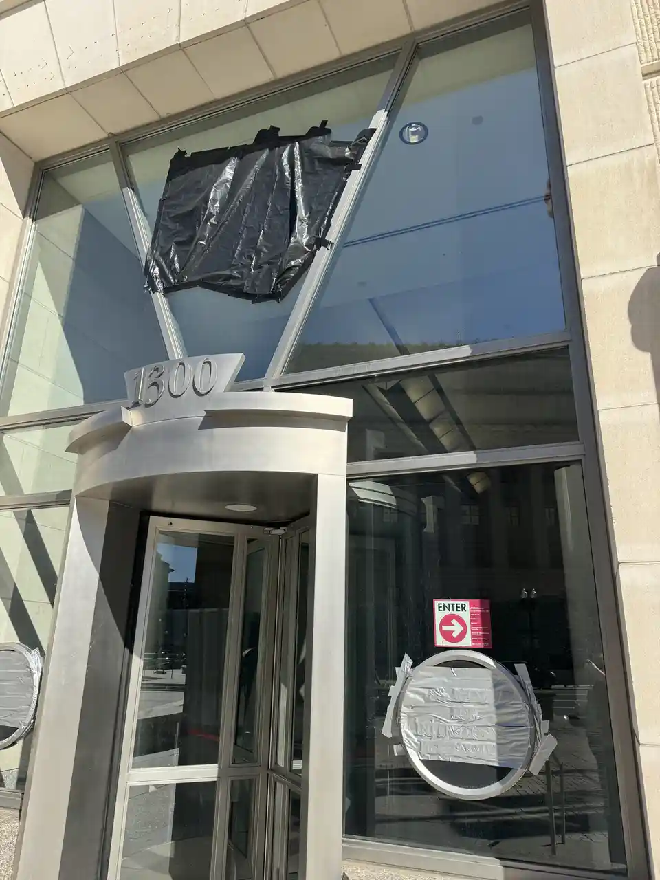 A modern building entrance with a revolving door marked '1300' has its windows covered with black plastic and silver tape. A…