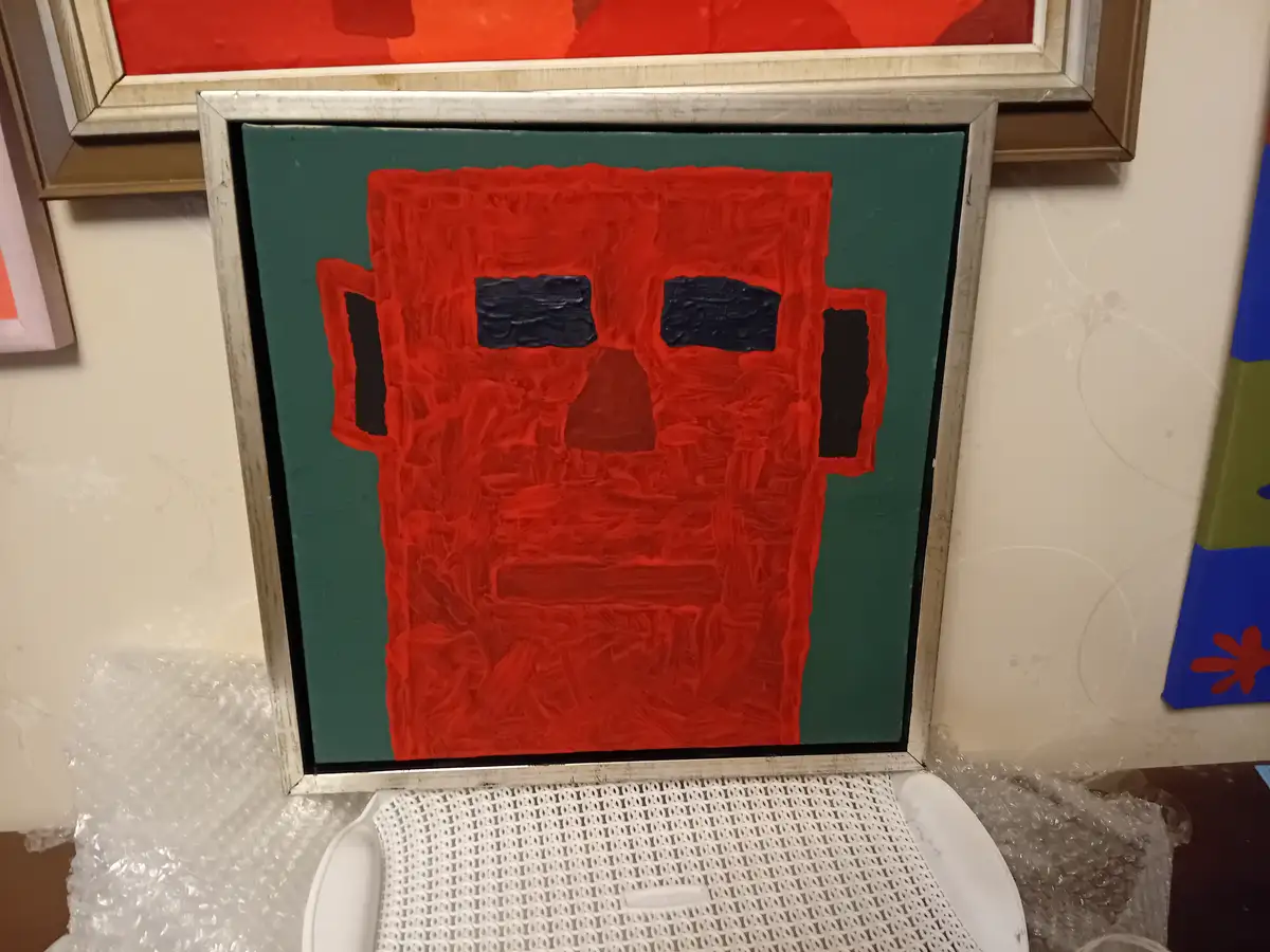 A framed painting of a red, blocky, abstract face with black eyes and mouth sits on a white plastic chair, surrounded by…
