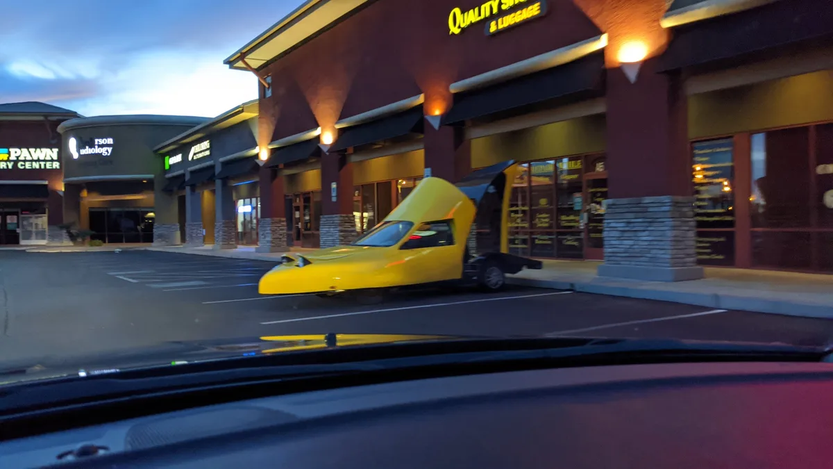 A bright yellow, futuristic-looking car with a shoe-like shape is parked in front of a 'Quality Storage & Luggage' store at…