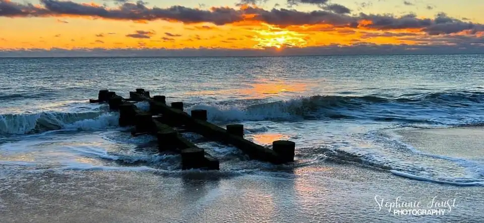A scenic photograph of a sunset over the ocean with waves crashing against a broken wooden pier extending into the water…