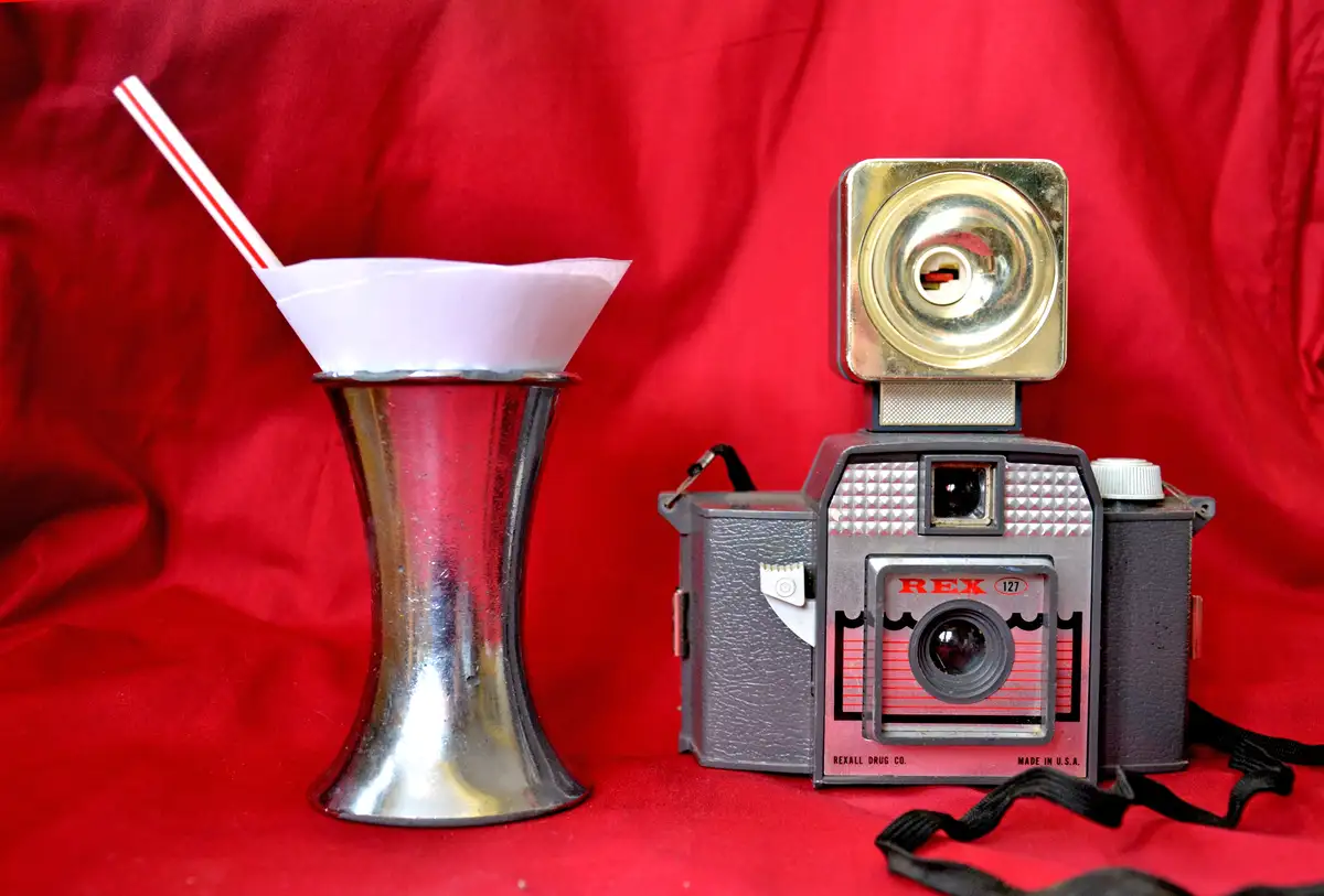 A vintage REX 127 camera sits next to a silver cocktail shaker with a paper cone and a red-and-white striped straw, both…