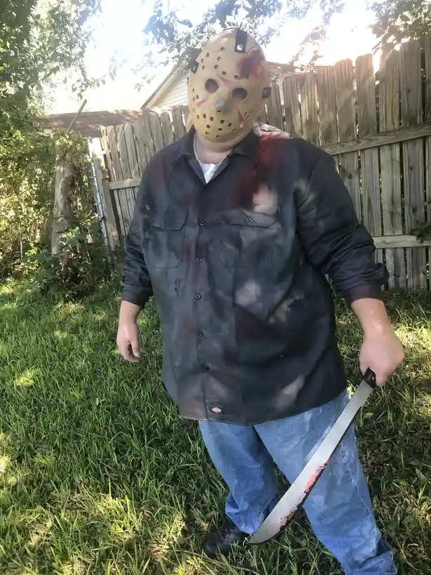 A person stands in a grassy backyard wearing a Jason Voorhees hockey mask with fake blood, a stained dark shirt, and jeans…