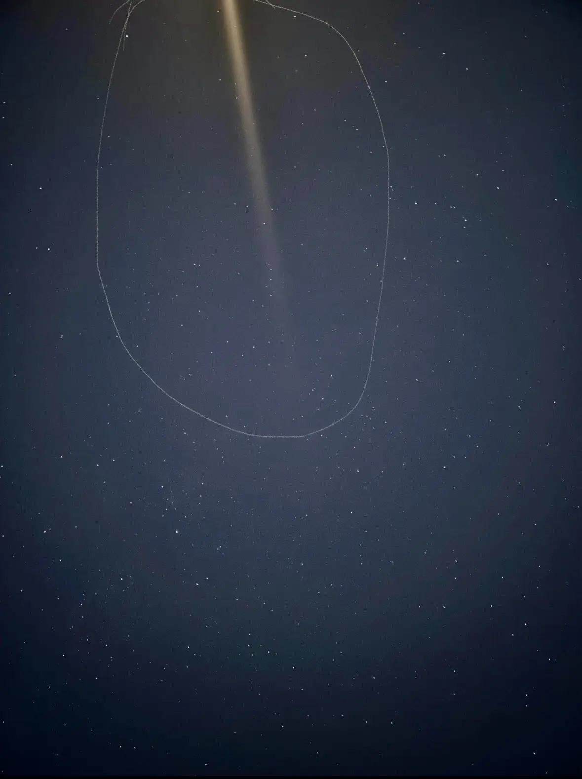 A dark night sky filled with stars, with a thin, hand-drawn white oval circling a bright vertical light beam, possibly from…