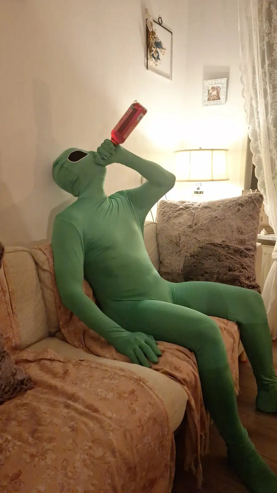A person in a full-body green alien costume sits on a patterned couch in a warmly lit living room, tilting their head back…