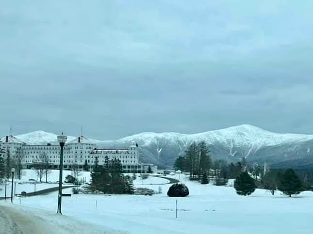 a snowy landscape featuring a large white hotel building in the foreground with snow-covered mountains in the background…
