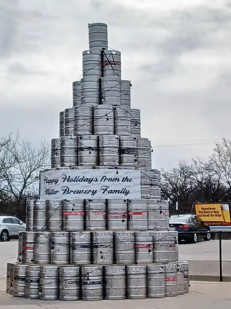 A large, pyramid-shaped structure made of stacked silver beer kegs stands outdoors under an overcast sky. A white banner…