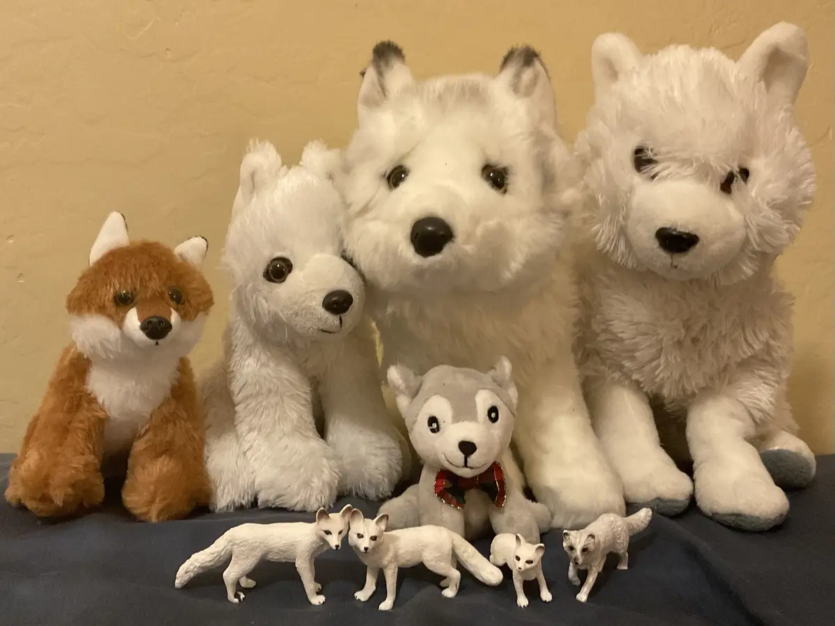 A collection of plush and figurine animals, including a brown fox, several large white wolves, a small grey husky with a…