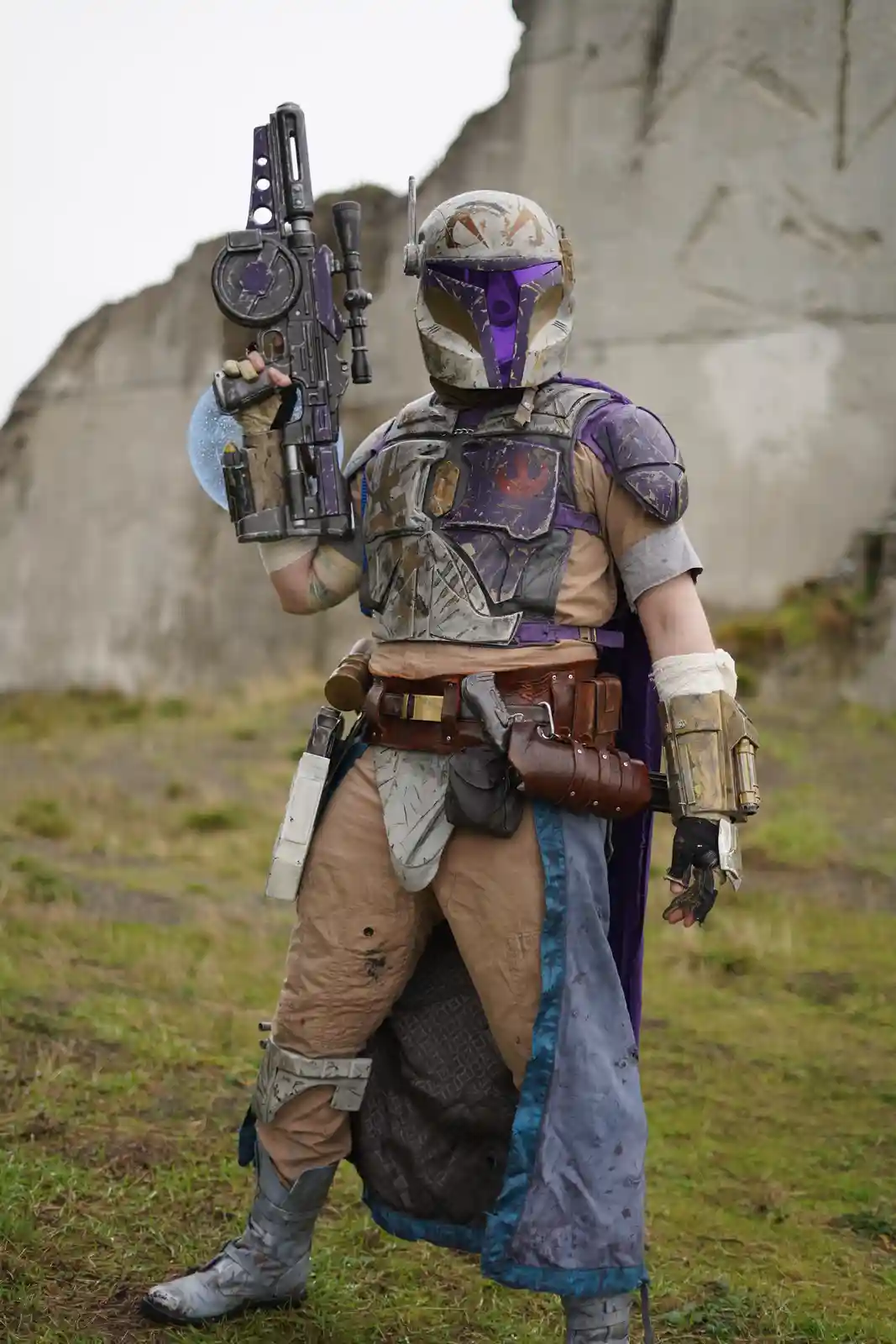 A person in a detailed, weathered Mandalorian-style costume with purple and silver armor, holding a large blaster rifle…