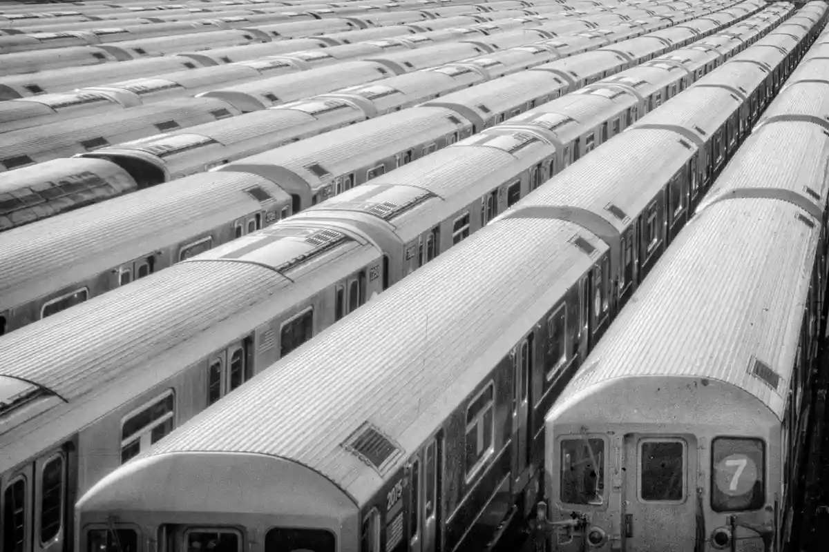 A black-and-white photo shows dozens of identical subway train cars parked in neat, parallel rows, stretching into the…