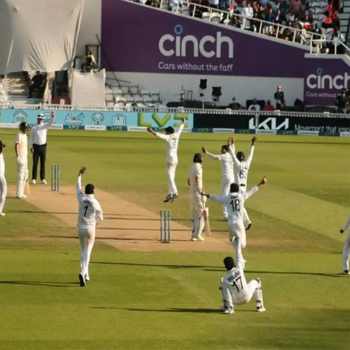 A group of cricket players in white uniforms celebrate enthusiastically on a green field, with some jumping and raising…