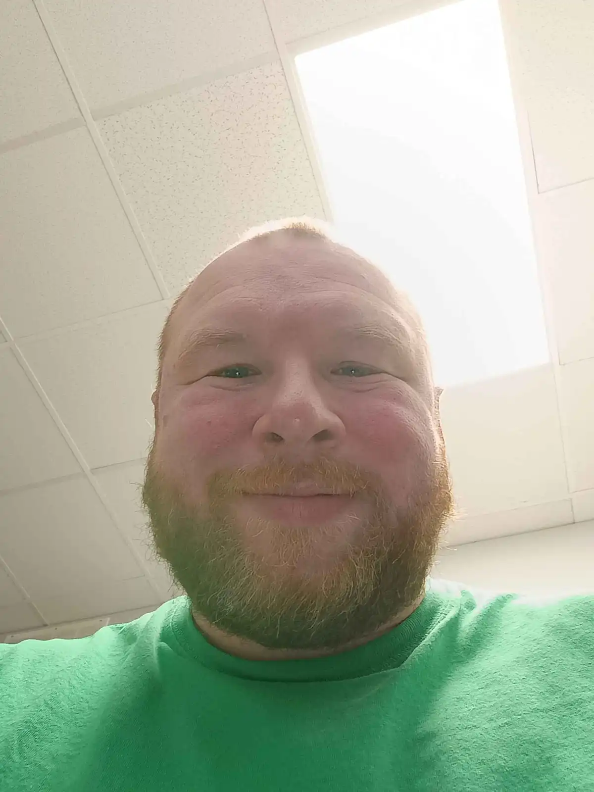 A selfie of a man with a reddish-brown beard and a green t-shirt, taken indoors with bright overhead lighting. He is smiling…