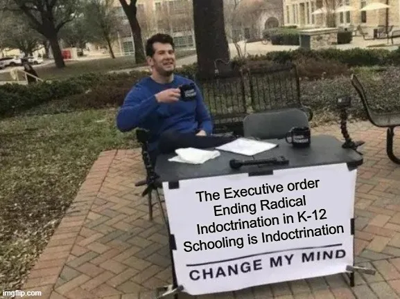 A man in a blue sweatshirt sits at a makeshift outdoor desk with a sign that reads 'The Executive order Ending Radical…