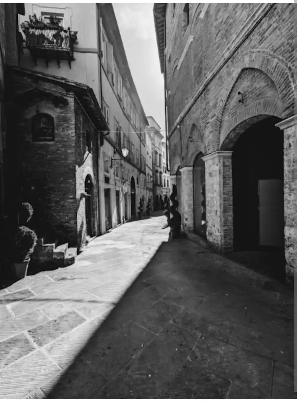 A black-and-white photo of a narrow, sunlit European alleyway with historic brick buildings and arched doorways. A person is…