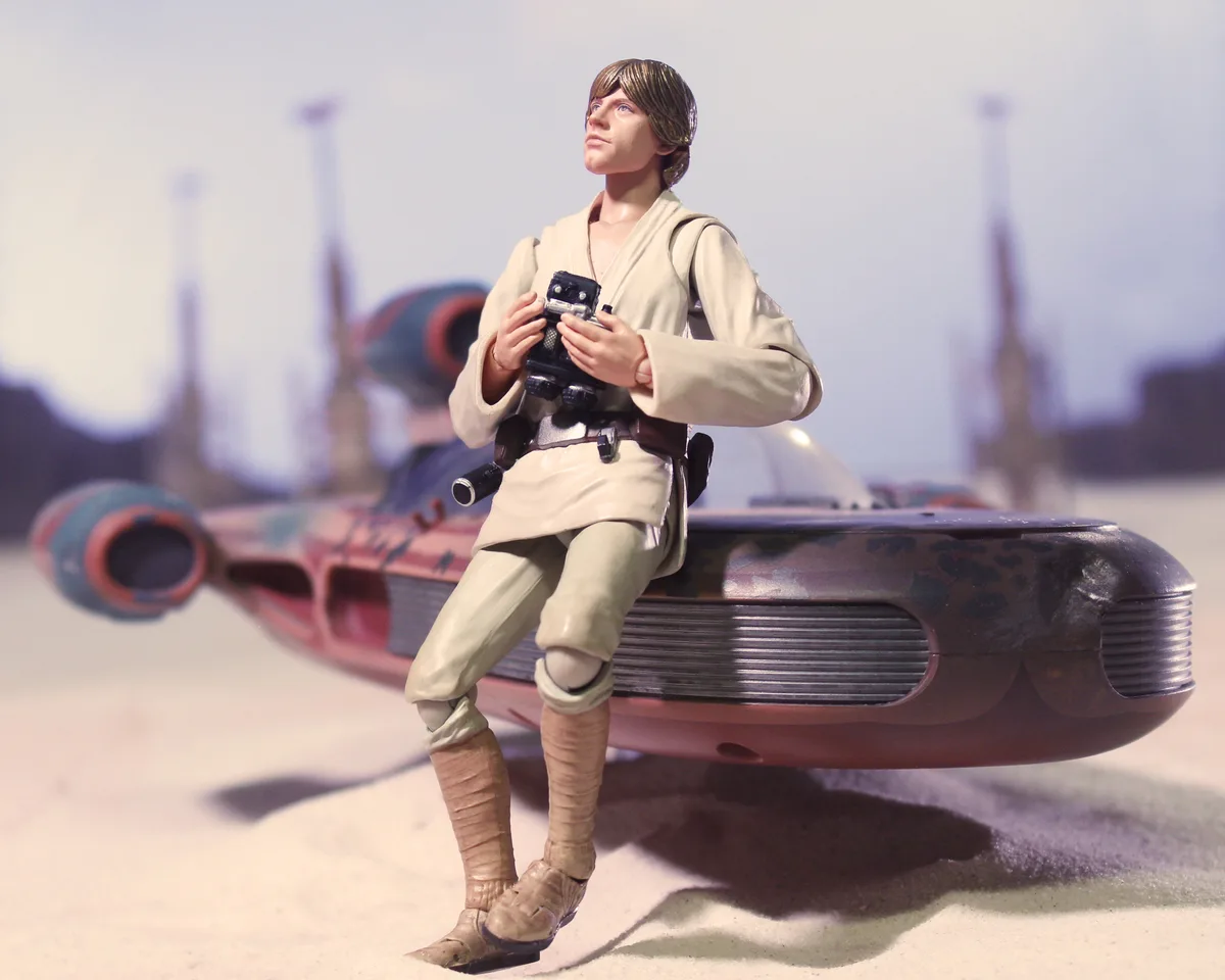 A Star Wars action figure of Luke Skywalker sits on the edge of a red and blue spaceship, holding a camera, against a desert…