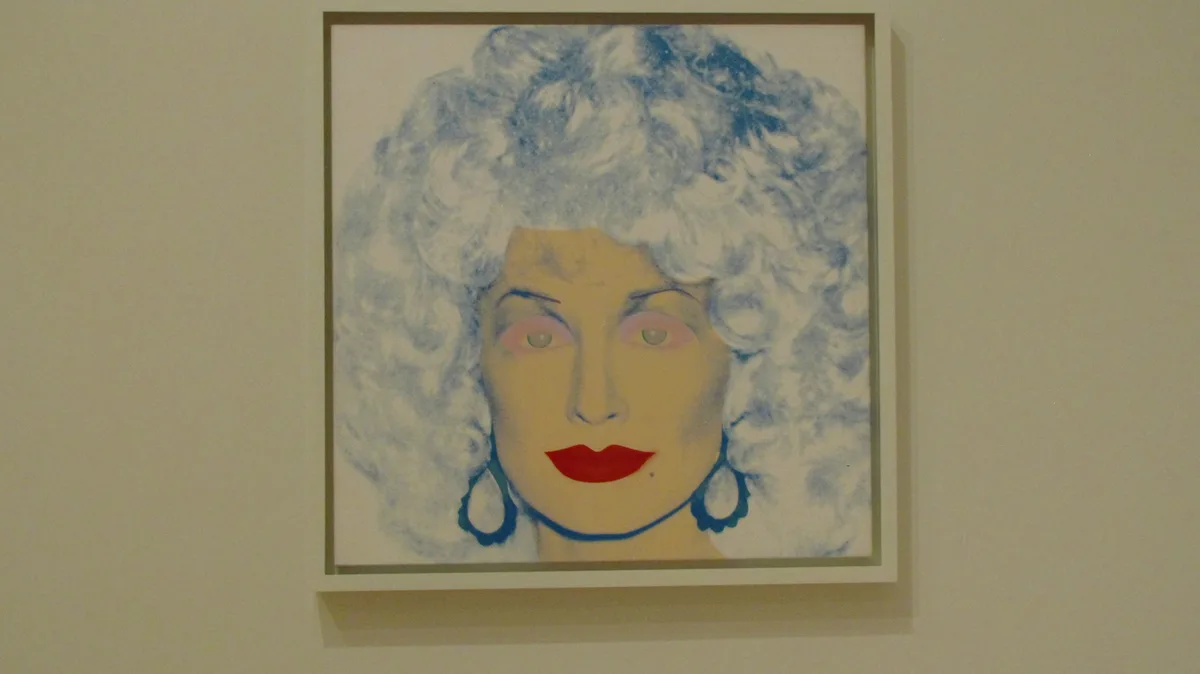 A framed painting of a woman with a large blue and white curly wig, pale skin, red lips, and large dangling earrings hangs…