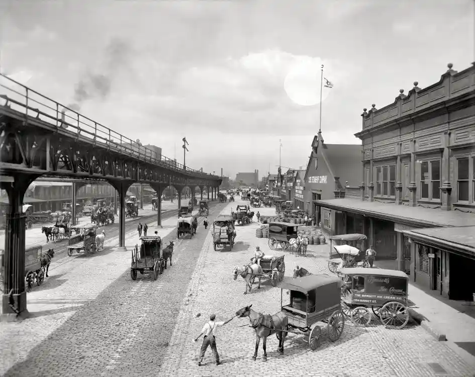 a black-and-white historical photograph of a busy 19th-century cobblestone street with horse-drawn carriages, pedestrians…
